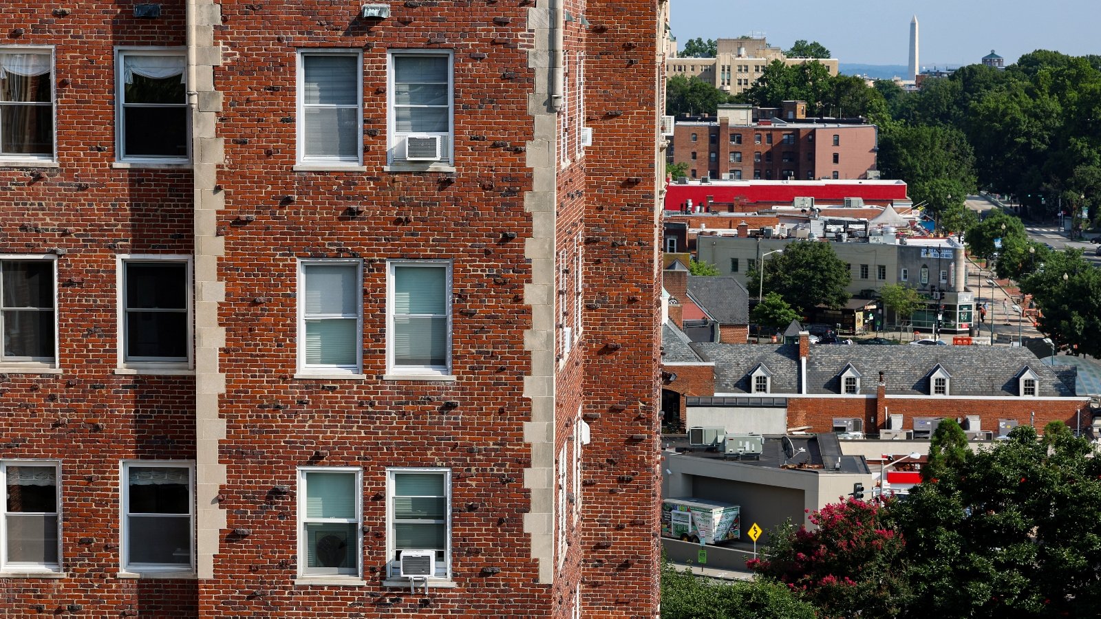 A large brick apartment building with AC window units stands in front of the DC skyline with the Washington monument in the background.